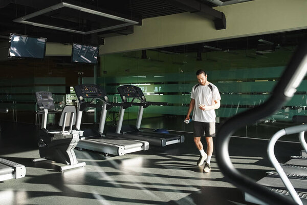 A man in active wear stands in a gym next to a row of treadmills.