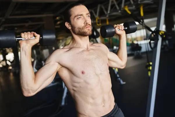 stock image A muscular man without a shirt holding two dumbbells in a gym, showcasing his workout routine and physical strength.