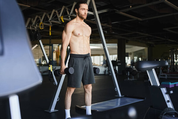 A shirtless man with sculpted muscles stands confidently with dumbbells in a gym, focused on his workout.