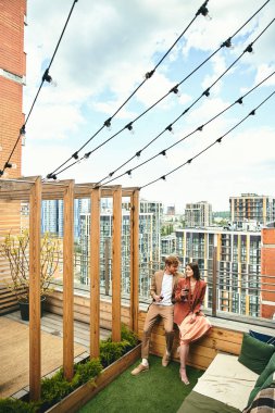 A man and woman sit closely on a rooftop, embracing each other as they enjoy the city skyline view below them clipart