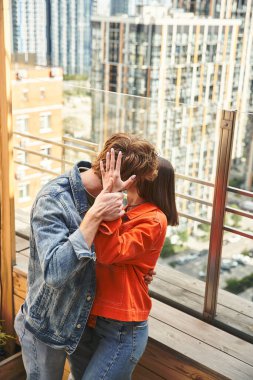 A man and a woman share a tender kiss on a balcony overlooking a cityscape, their bodies pressed together in an intimate embrace clipart