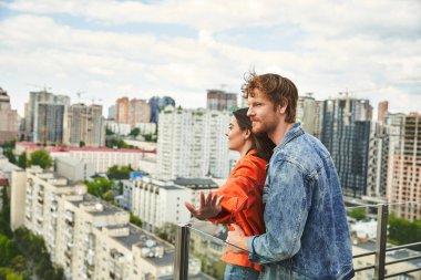 Bir adam ve bir kadın bir balkonda dikilip gökyüzünün altındaki geniş şehre bakıyorlar.