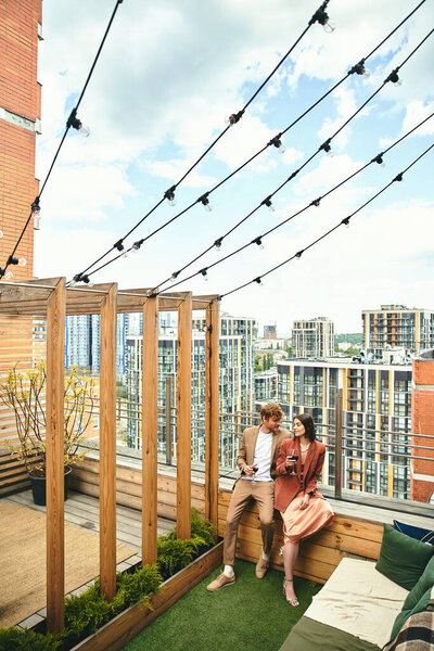 A man and woman sit closely on a rooftop, embracing each other as they enjoy the city skyline view below them