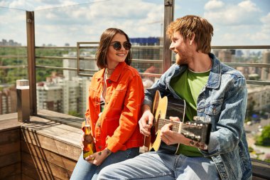 Bir adam ve bir kadın bir binanın tepesinde oturuyor, aşağıdaki şehre bakıyor, birlikte sakin bir anın tadını çıkarıyorlar.
