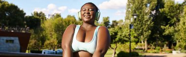 Kıvrımlı bir Afrikalı Amerikalı kadın, parkta güvenle duruyor, kulaklıkla müzik dinliyor ve vücudunun pozitifliğini kucaklıyor..