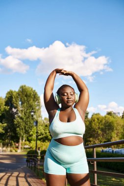 Afro-Amerikalı bir kadın açık havada mavi bir spor sütyeni ve uyumlu şortla, vücut pozitifliği ve özgüveniyle coşkuyla egzersiz yapıyor..