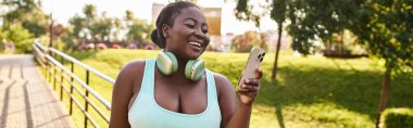 An African American woman joyfully listens to music on her cell phone while wearing headphones outdoors. clipart