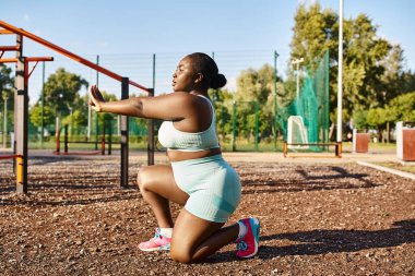 Spor giyim sektöründe bir Afrikalı Amerikalı kadın oyun parkının önünde çömeliyor, vücut pozitifliği ve gücü somutlaştırıyor..