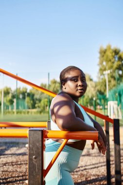 Spor kıyafetleri içinde, vücudu pozitifliğini gösteren Afro-Amerikalı bir kadın, doğa ile çevrili bir bankta oturuyor..