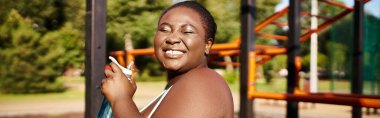 Spor giyimli Afrikalı bir Amerikalı kadın oyun parkının önünde kendinden emin bir şekilde duruyor, vücut pozitifliği ve gücü somutlaştırıyor..