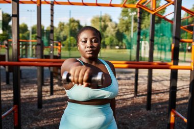 Mavi spor giysili kıvrımlı Afrikalı Amerikalı bir kadın kendinden emin bir şekilde elinde metal bir dambıl tutuyor..