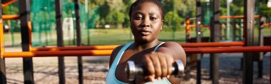 Mavi spor giysili kıvrımlı Afrikalı Amerikalı bir kadın halterini elinde tutuyor, zarafet ve güç saçıyor..