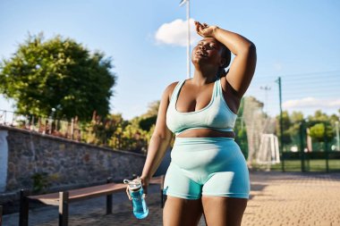 African American woman in sports bra and shorts holding a water bottle, staying hydrated during her outdoor workout. clipart