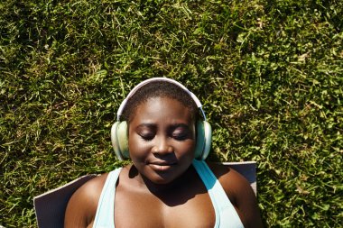 Afro-Amerikalı bir kadın spor kıyafetleriyle doğada dinleniyor, otların üzerinde yatarken kulaklıkla müzik dinliyor..