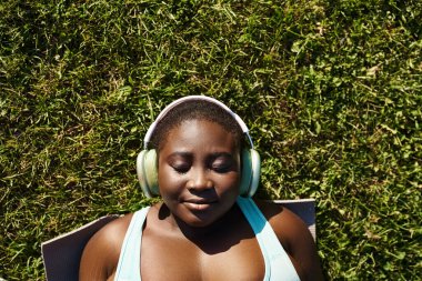 An African American woman in sportswear relaxes in nature, listening to music through headphones while laying in the grass. clipart