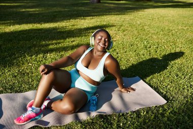 Spor kıyafetli kıvrımlı Afrikalı Amerikalı bir kadın yoga minderinde zarif bir şekilde oturmuş açık havada farkındalık egzersizi yapıyor..