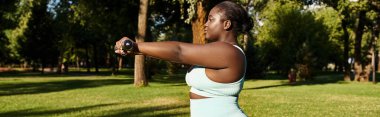 An African American woman in a white tank top confidently holds a black dumbbells outdoors, embodying body positivity and strength. clipart