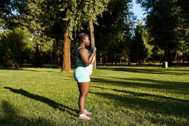 A curvy African American woman in sportswear stands gracefully in the grass, holding dumbbells clipart