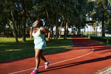 Spor giyim sektöründe Afro-Amerikalı bir kadın enerjik bir şekilde zengin bir park ortamında koşuyor, pozitifliği ve kendine güveni somutlaştırıyor..