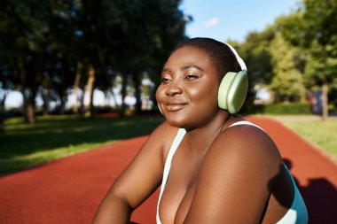 Spor kıyafetleri içinde, kulaklık takmış, müziğe dalmış, açık havada vücut pozitifliğini somutlaştıran Afrikalı bir Amerikalı kadın oturuyor..
