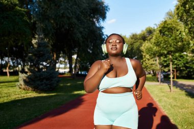 Spor kıyafetleriyle koşan, kulaklık takan ve dışarıda egzersiz yapan kıvrımlı Afrikalı Amerikalı bir kadın..