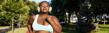Spor kıyafetleri içinde, vücudunun pozitifliğini kucaklayan Afrikalı Amerikalı bir kadın, kulaklık takarak parkta koşuyor, doğa seslerini açıyor..