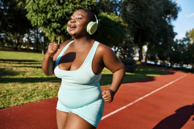 Spor kıyafetleri içinde bir Afrikalı Amerikalı kadın bir pistte koşarken kulaklıkla müzik dinliyor, vücut pozitifliğini somutlaştırıyor..