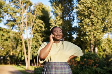 Artı, normal giyinmiş Afrikalı Amerikalı bir kadın, vücut pozitifliğini kucaklıyor, güneşli bir günde parkta cep telefonuyla sohbet ediyor..