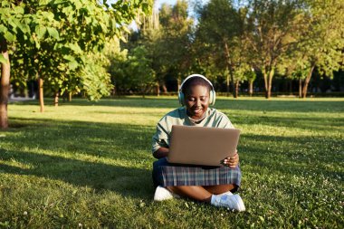 Büyük boy Afro-Amerikan bir kadın gündelik giysiler içinde çimenlerde dizüstü bilgisayarla oturuyor ve yazın açık havada verimli bir çalışma yapıyor..