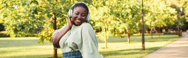Büyük boy Afro-Amerikan kadın günlük giysiler içinde, vücut pozitifliği sergiliyor, kulaklıkla müzik dinliyor.