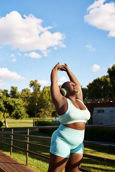 stock image An African American woman in a blue top and shorts energetically stretches her arms outdoors, embodying body positivity.