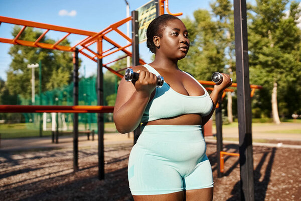 An African American woman in sportswear proudly lifts a dumbbell in a peaceful park setting.