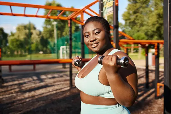 一名身穿运动服的非裔美国妇女自信地在茂密的公园里带着哑铃进行运动 拥抱身体的积极向上 — 图库照片