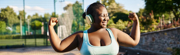 An African American woman in a sports bra top flexes her muscles confidently outdoors, showcasing body positivity and strength.