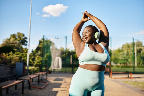 Curvy African American woman in sports bra top stretches her arms outdoors, promoting body positivity and fitness.