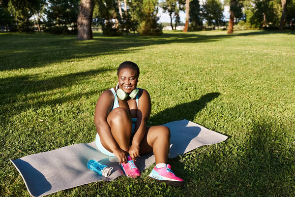 Игривая афроамериканка в спортивной одежде, сидящая на полотенце, наслаждающаяся природой в мирной и спокойной обстановке.