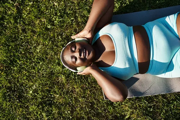 スポーツウェアのアフリカ系アメリカ人女性が緑色の草のマットでヨガを実践し 体のポジティブさを受け入れる — ストック写真