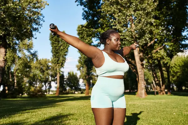 stock image A curvy African American woman in blue sports bra and shorts holds a black dumbbells while exercising outdoors.