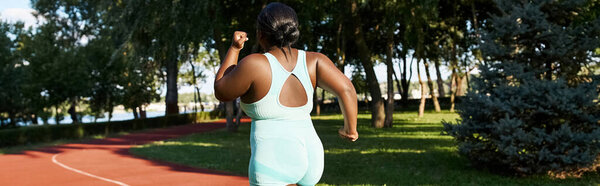A curvy African American woman in sportswear runs along a red track on a sunny day