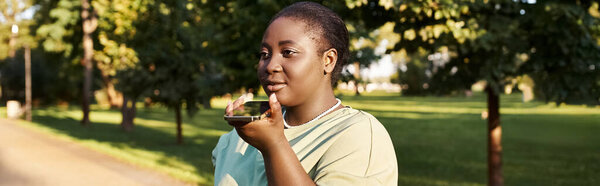 A young woman, plus size and African American, holds a smartphone in her hand outdoors in the summer, embracing body positivity.