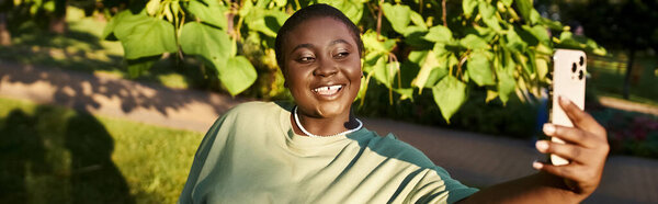 Plus-size African American woman captures a carefree moment, snapping a selfie outdoors in casual attire during summer.
