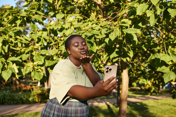 Плюс размер афроамериканка стоит под деревом, держа сотовый телефон в солнечный день.
