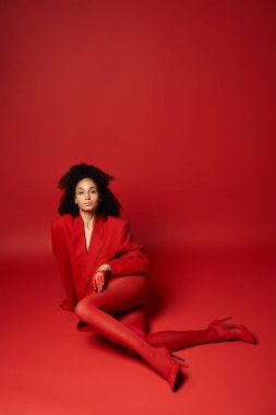 Genç bir kadın zarif bir şekilde yerde oturuyor. Parlak kırmızı bir ceket ve tayt giyiyor. Stüdyoda canlı bir arka plana karşı..