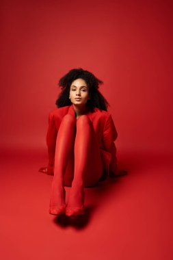 Kırmızı ceketli ve taytlı genç bir kadın canlı bir stüdyo ortamında yerde oturuyor..