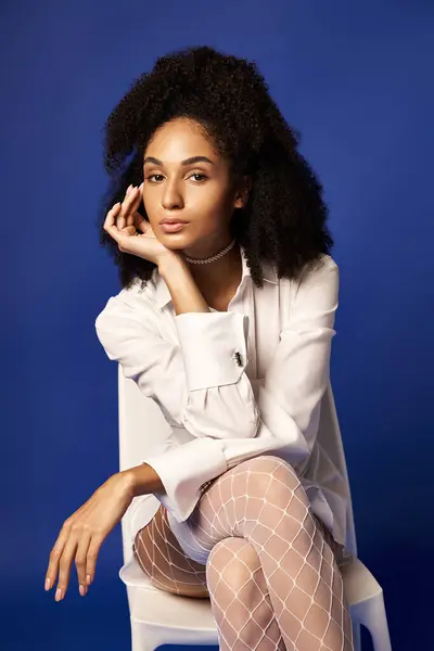 stock image A young woman in a white dress and stockings sits gracefully atop a white chair against a blue studio background.