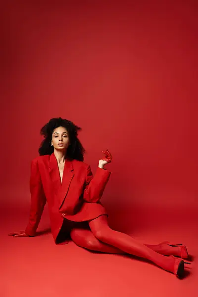 stock image A young woman in a vibrant red suit sitting gracefully on the floor in a studio setting.