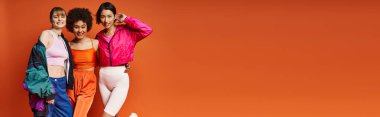 Üç farklı kadın, Kafkasyalı, Asyalı ve Afrikalı Amerikalı, turuncu arka planda bir stüdyoda yan yana duruyorlar..