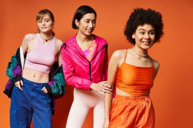 Beyaz, Asyalı ve Afro-Amerikan kökenli üç kadın turuncu arka planlı bir stüdyoda zarifçe duruyorlar..