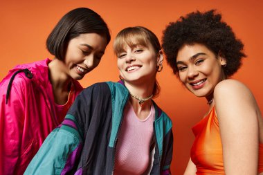Farklı etnik kökenlere sahip üç güzel kadın turuncu bir arkaplanın önünde yan yana duruyor..