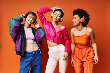 Aralarında Kafkas, Asyalı ve Afrikalı Amerikalıların da bulunduğu bir grup genç kadın, turuncu bir stüdyo geçmişine karşı birlikte duruyorlar..